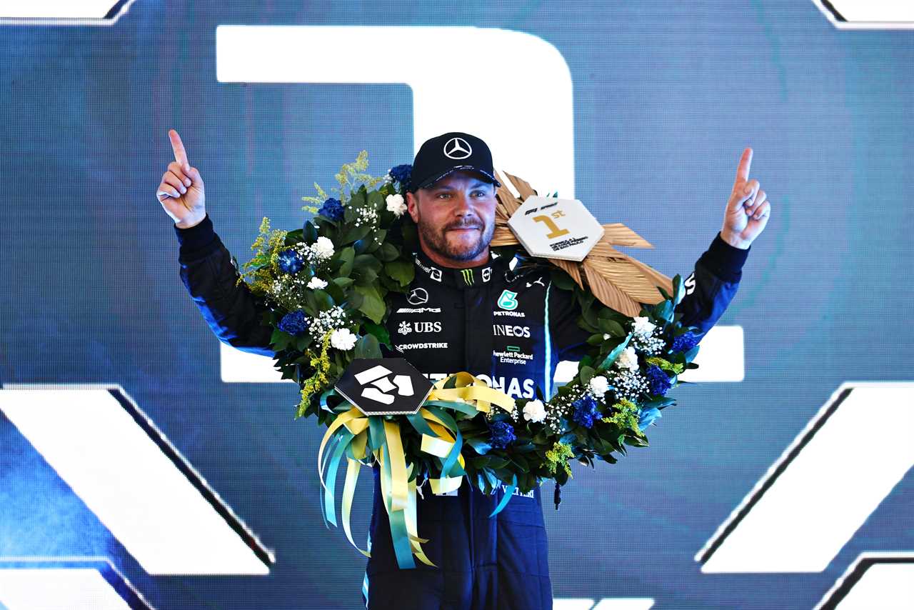 Valtteri Bottas wears a flower garland with a 1 around his neck and holds his fingers up to the sky